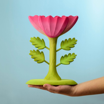 Hand holding a wooden large pink flower with a green stem and leaves, against a blue background.
