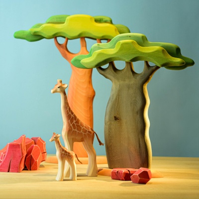 Wooden giraffe family toys next to baobab trees and rocky terrain.