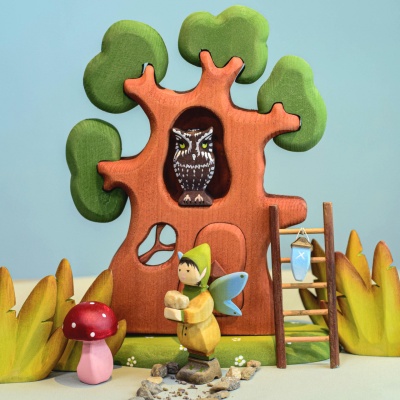 Wooden winged elf figure standing in front of a whimsical wooden tree house, with an owl in the tree and a ladder leaning against it. The elf is surrounded by small rocks and a red mushroom.