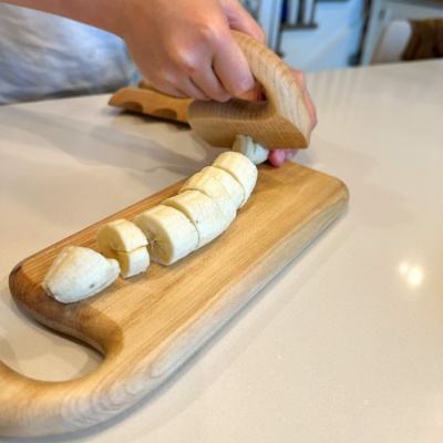 Child using wooden chopper on a cutting board to slice a banana