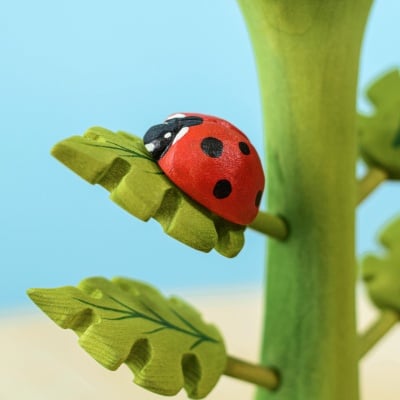 Vivid red wooden ladybug toy with black spots perched on a green leaf attached to a wooden stem, against a soft blue background.