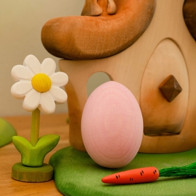 Pink wooden Easter egg next to a white wooden daisy and a toy carrot near a handcrafted wooden fairy house