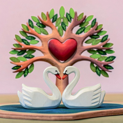 Two swans standing together in front of a tree with a heart carved into its trunk. The swans are white with orange beaks and black feet. The tree is brown and has green leaves.
