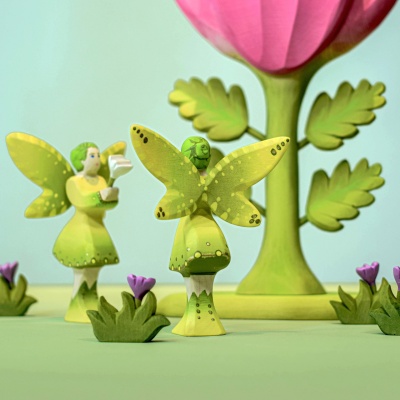 Wooden large pink flower surrounded by multiple green-winged fairy figures, some standing and one on top of the flower, in a whimsical setting with small green plants with purple flowers.