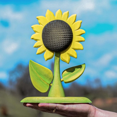 A sunflower wooden sculpture held in a hand, with bright yellow petals and a textured brown center, set against a clear blue sky.