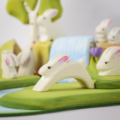 White wooden Easter bunny figurine running on a green base, with pink inner ears, surrounded by trees and a purple tulip.