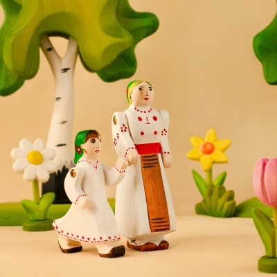 Traditional Romanian wooden figures of a woman and child walking past a yellow daffodil and spring flowers