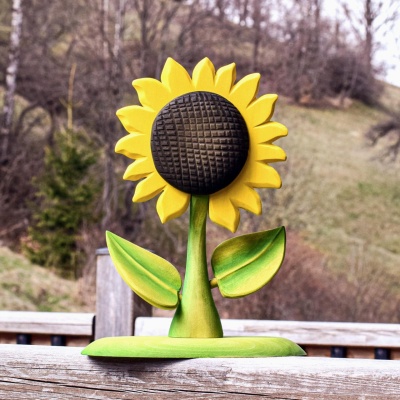 A handcrafted wooden sunflower toy stands on a wooden railing with a natural blurred forest background.