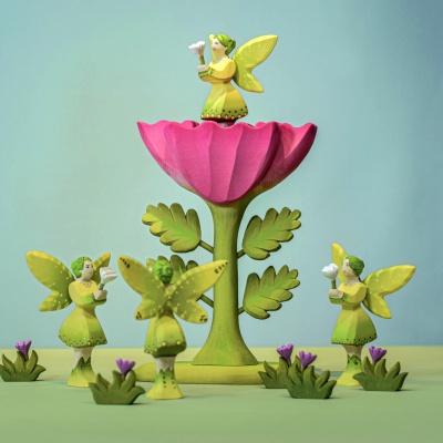 Wooden large pink flower surrounded by multiple green-winged fairy figures, some standing and one on top of the flower, in a whimsical setting with small green plants.