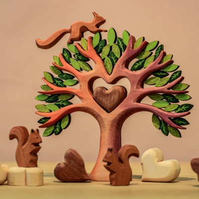 A handcrafted wooden tree with a heart-shaped cutout in its trunk, surrounded by several small wooden squirrels and additional hearts. The tree and decorations are placed on a wooden table.