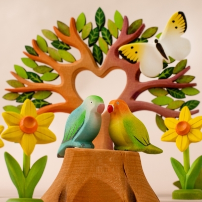 Two wooden toy birds resting on a tree stump surrounded by a heart-shaped tree and two yellow handcrafted daffodils