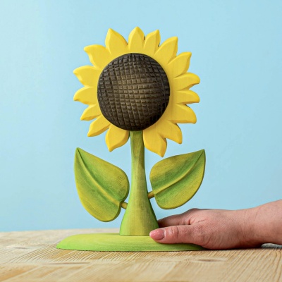 A wooden sunflower toy with yellow petals and a woven pattern center, standing on a wooden surface against a light blue background.