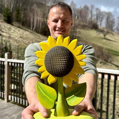 A person holding a large wooden sunflower decoration with vivid yellow petals, green leaves, and a detailed center against a rural backdrop.