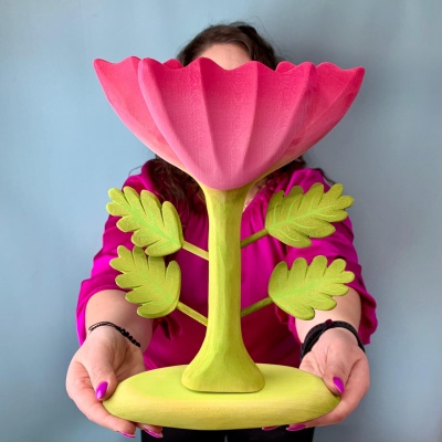 Person in a pink shirt holding a large wooden pink flower with a green stem and leaves, against a blue background.