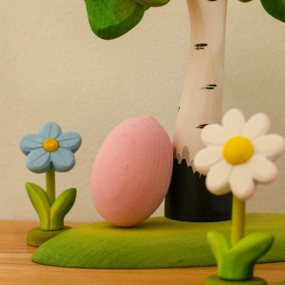 Pink wooden egg leaning against a toy birch tree next to blue and white wooden flowers on a green play base