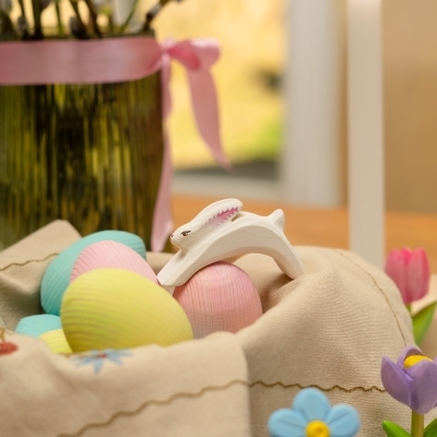 Child hands placing a pastel pink wooden Easter egg next to a fairy house and white bunny figures