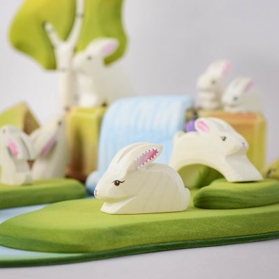 "White wooden Easter bunny figurine in a perching pose on a green base, with pink inner ears, surrounded by green trees and a blue waterfall background.