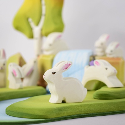 White wooden Easter bunny figurine in a sitting pose on a green base, with pink inner ears, surrounded by green trees and a blue waterfall background.