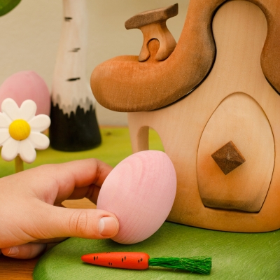 Childs hand holding a pink wooden Easter egg next to a handcrafted wooden fairy house