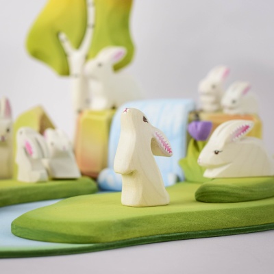 White wooden Easter bunny figurine in a curious pose on a green base, with pink inner ears, surrounded by green trees and a blue waterfall background.