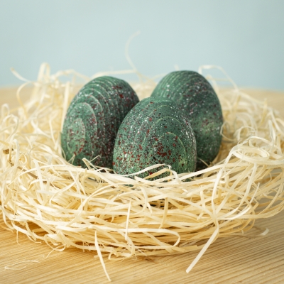 Three green speckled wooden dinosaur eggs resting in a straw nest on a wooden surface.