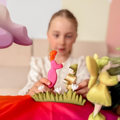 Child playing with a wooden winged elf figure and another wooden figure with red hair and a pink dress. The scene includes grass and flowers on a colorful table.