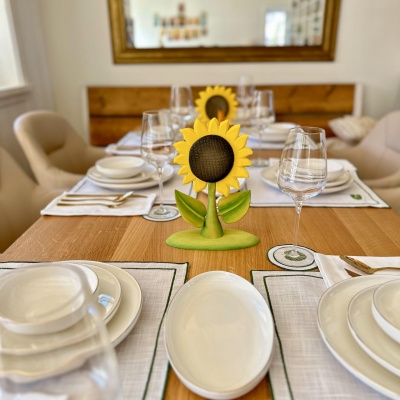 Close-up of a wooden sunflower table decoration, with yellow petals and green leaves, amidst a set dining table with white dishes.