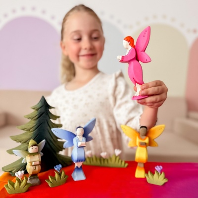 Smiling child holding a wooden fairy figure with pink wings and dress, with other wooden fairy figures in blue, yellow, and green on a colorful table.