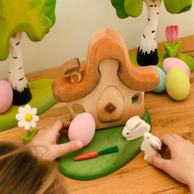 Child playing with a wooden fairy house, white toy bunnies, pastel eggs, and wooden trees