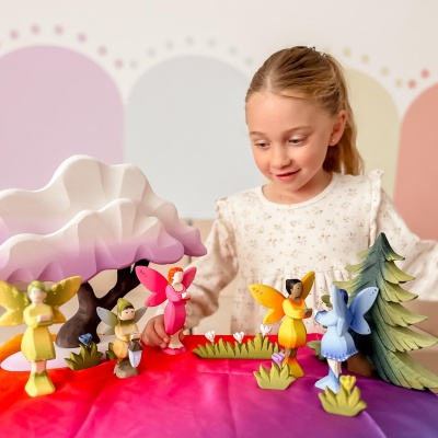 Child playing with multiple wooden fairy figures in various colors in a whimsical scene with trees and flowers on a colorful table.