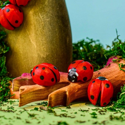 Multiple red wooden ladybug toys scattered on and around a wooden log, set against a green mossy backdrop, evoking a natural setting.