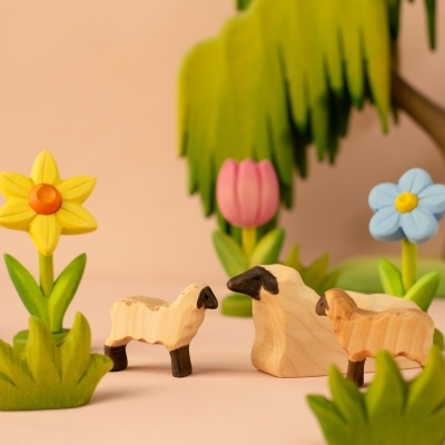 Handcrafted wooden sheep and lamb figures grazing near a yellow daffodil and blue spring flower