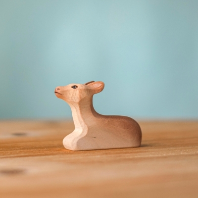 Handmade wooden Sitting Reindeer Calf figure with head raised, resting on a table against a solid blue background.