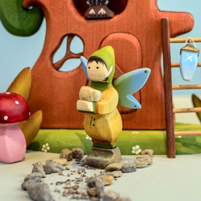 Wooden winged elf figure in a green hooded outfit with blue wings, holding a small stone, standing in front of a whimsical wooden tree house with a red mushroom nearby.