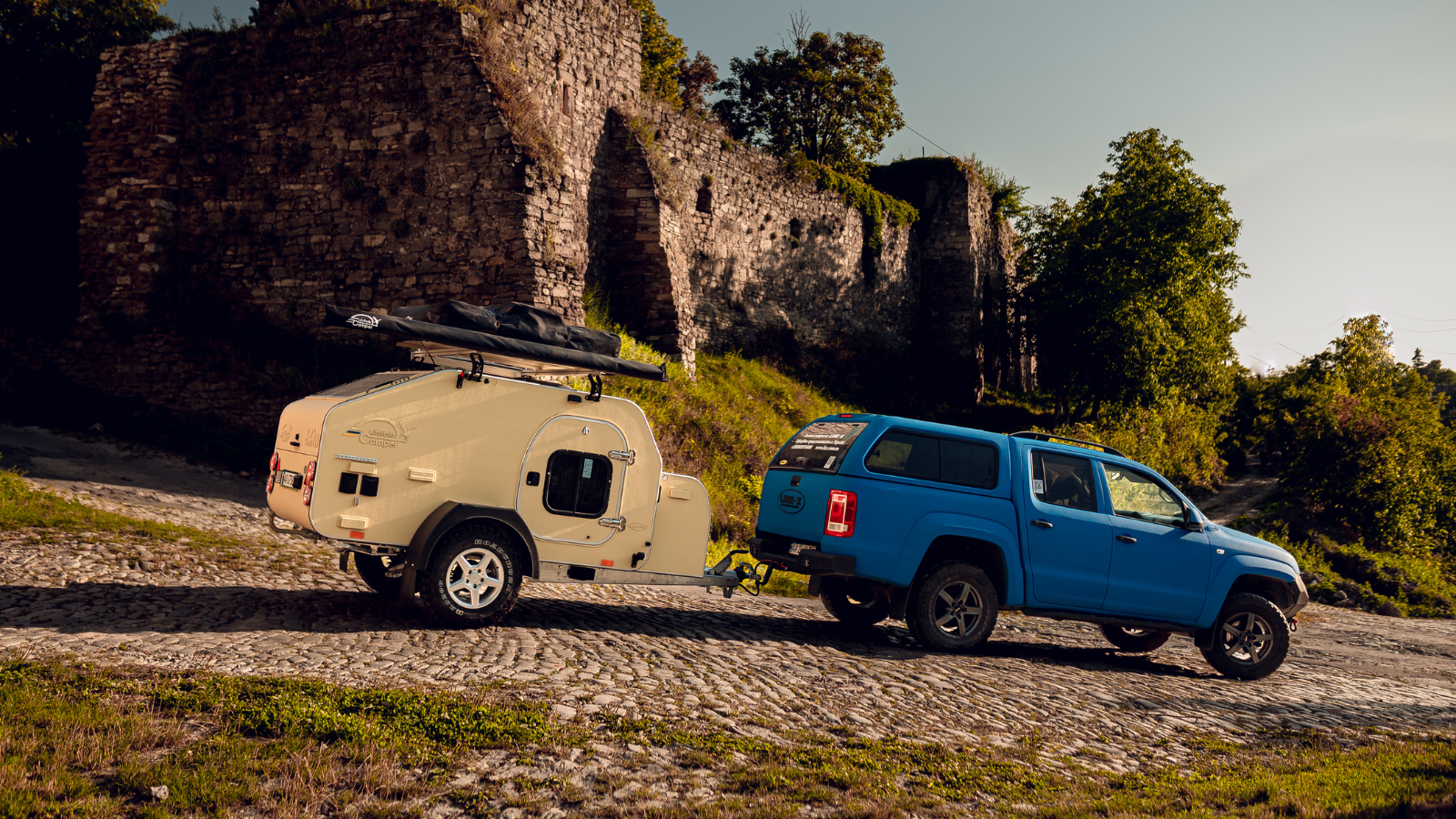 Rulotă teardrop deschisă la culoare, lângă un pick-up albastru