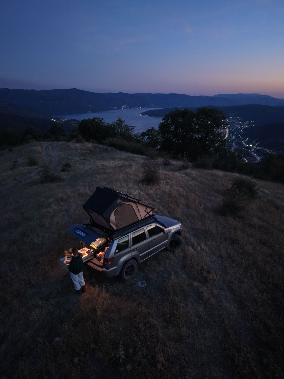 Vedere de sus, la apus: un 4×4 cu cortul de acoperiș deschis pe un deal, cu un om lângă mașină și apa în depărtare