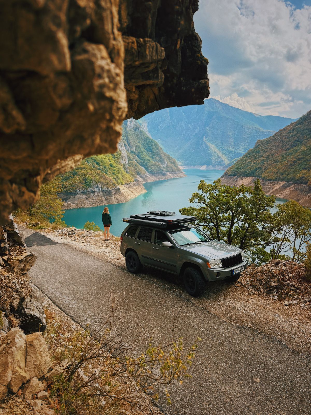 Un 4×4 pe un drum îngust sub o stâncă ieșită în afară, deasupra unui lac turcoaz între munți, cu un om în dreapta