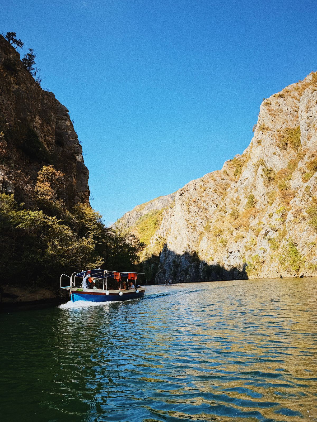 O barcă pe apa albastră a Canionului Matka, între pereți de stâncă, sub cer senin