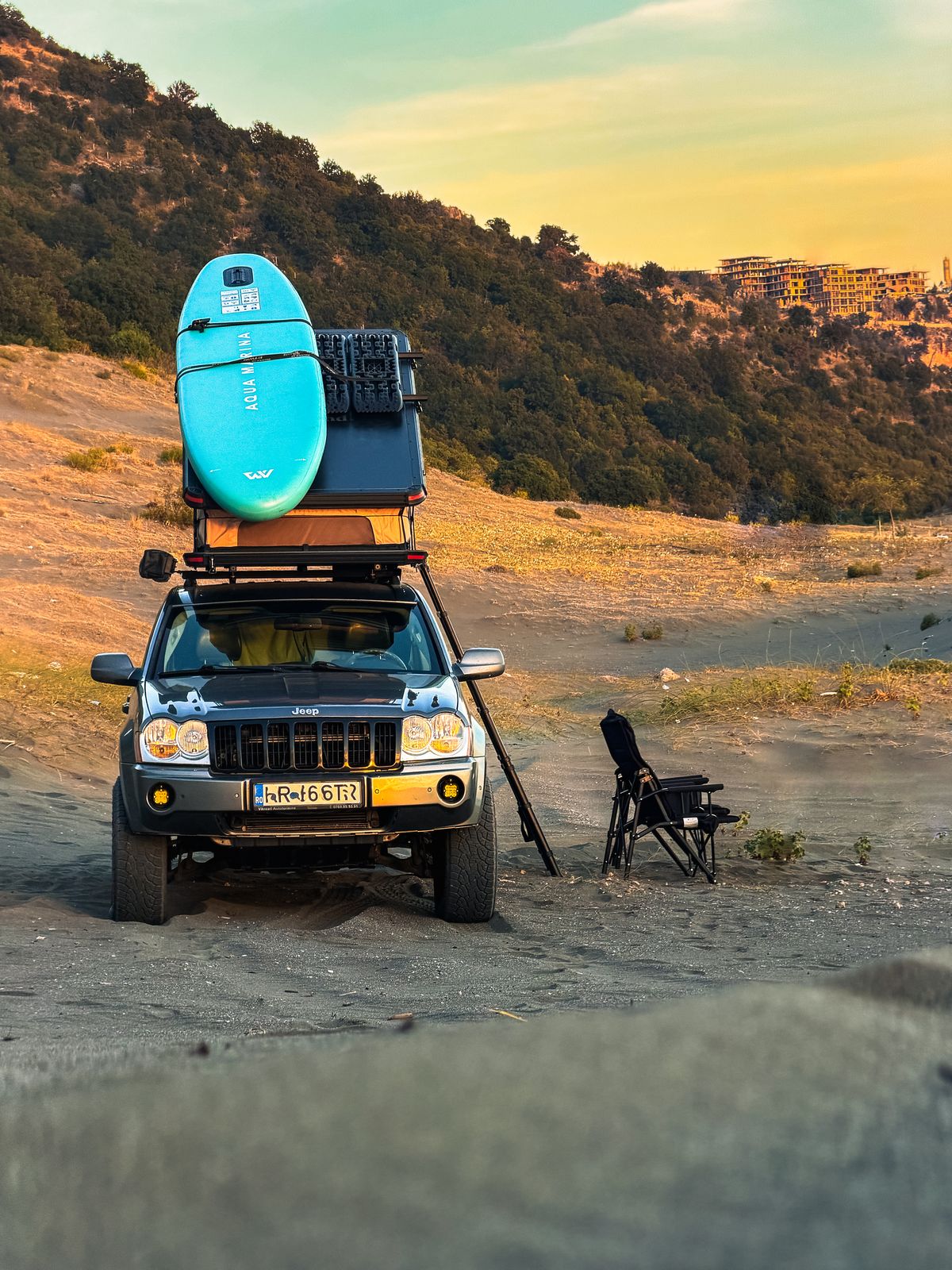 Un 4×4 cu o placă SUP turcoaz și cutii pe portbagajul de plafon, lângă un scaun de camping, în lumina apusului