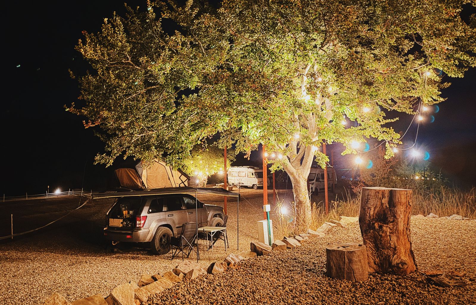 Un camping noaptea, cu un copac mare luminat de ghirlande, o mașină și un cort în fundal