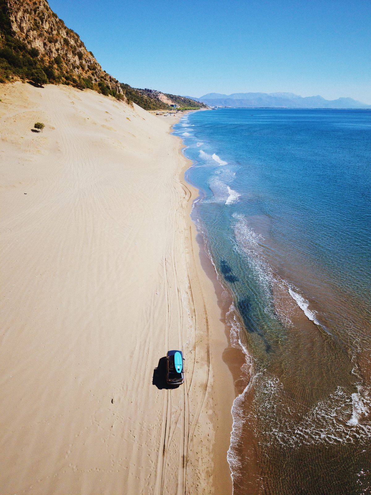 Vedere de sus a unei plaje lungi cu nisip, cu marea pe dreapta și un 4×4 mic pe nisip