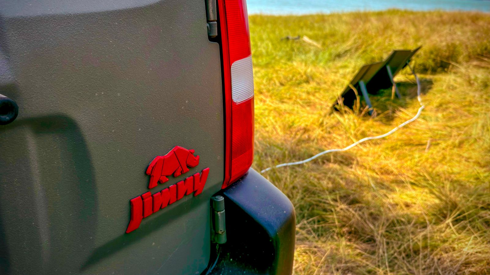 Spatele unui Suzuki Jimny verde lângă un lac, cu un panou solar portabil pe iarbă