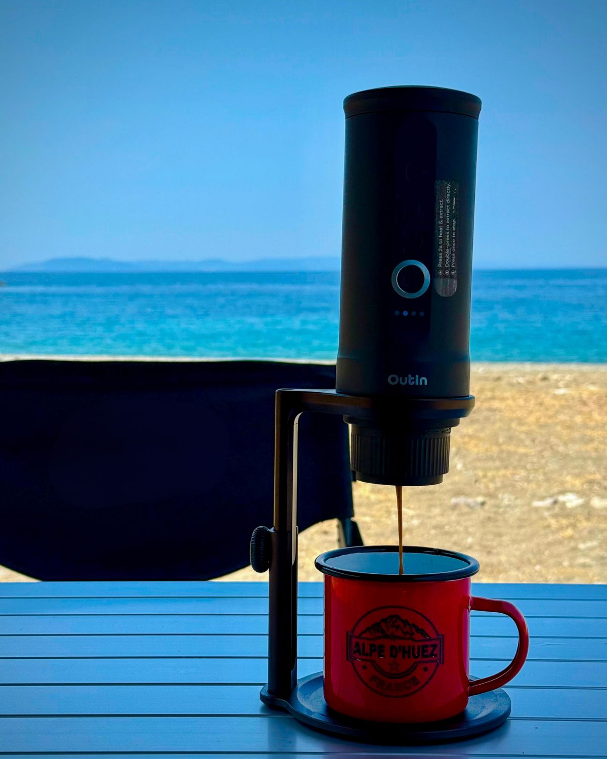 Espressor portabil care pregătește cafea într-o cană roșie, pe o masă de camping, cu marea în fundal