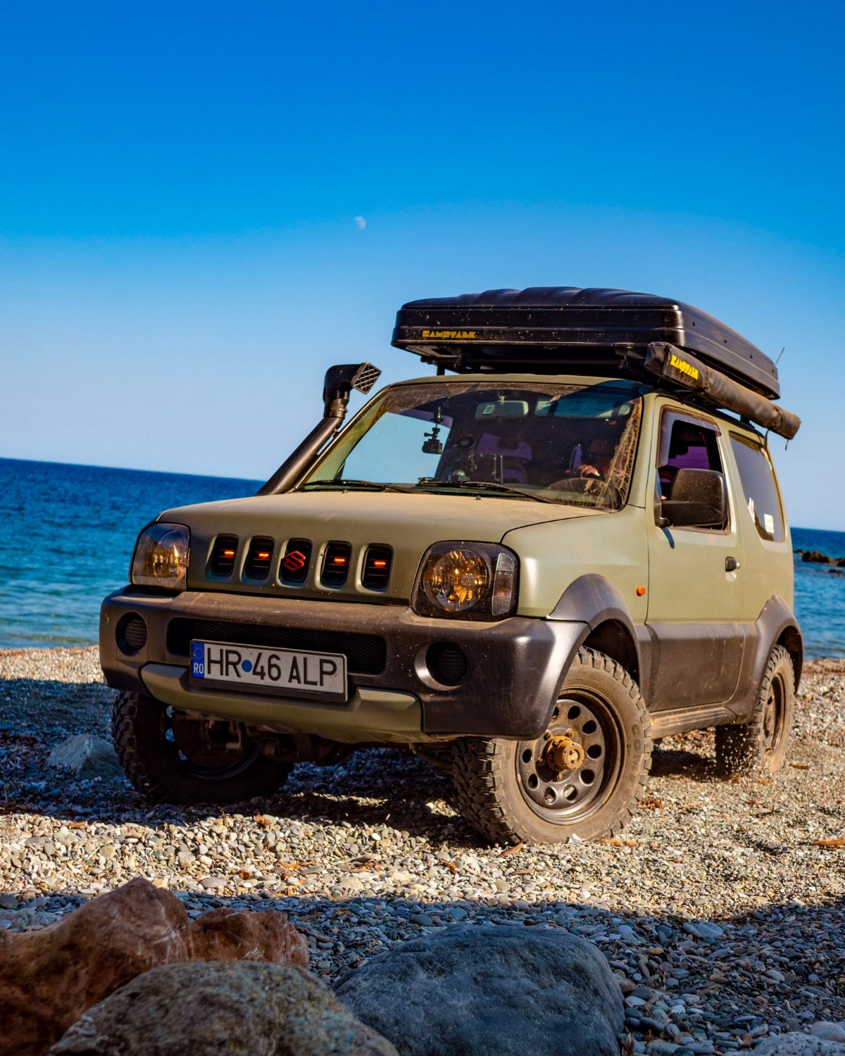 Suzuki Jimny cu cort de plafon închis, parcat pe o plajă cu pietriș lângă mare
