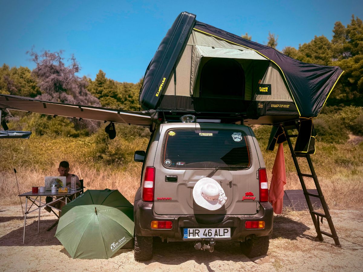 Suzuki Jimny văzut din spate, cu cortul de plafon deschis, scara coborâtă și o masă de camping sub marchiză