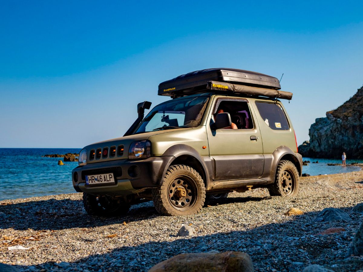 Suzuki Jimny pe o plajă cu pietriș, cu cortul de plafon cu carcasă rigidă închis