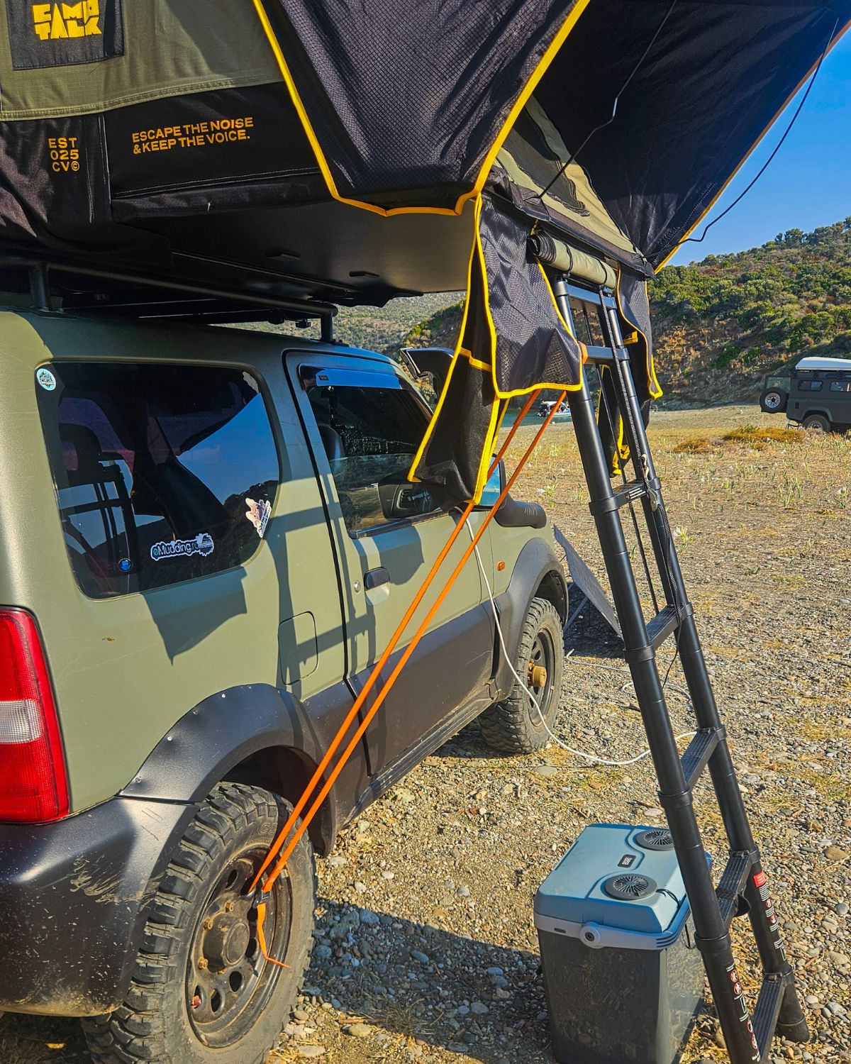Cort de plafon CampTalk deschis pe un Jimny, cu scara coborâtă și o chingă portocalie legată de roată