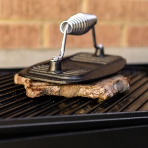 Presa de carne din fontă în uz, pe grătar, cu o bucată de carne.