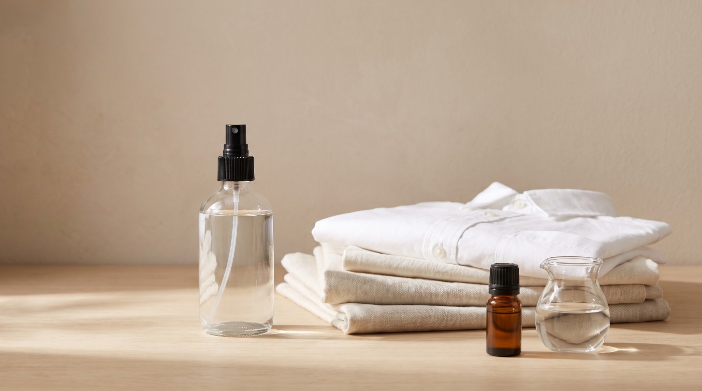 Sticlă de spray cu parfum de rufe lângă o cămașă albă călcată și șervete de in împăturite
