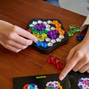 Joc IQ Deluxe Pentagon de la Smart Games cu tablă de joc pentagonală, 11 piese puzzle translucide colorate și broșură cu 120 de provocări logice progresive.
